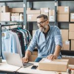 Older man working on computer in stock room with boxes and shirts around him